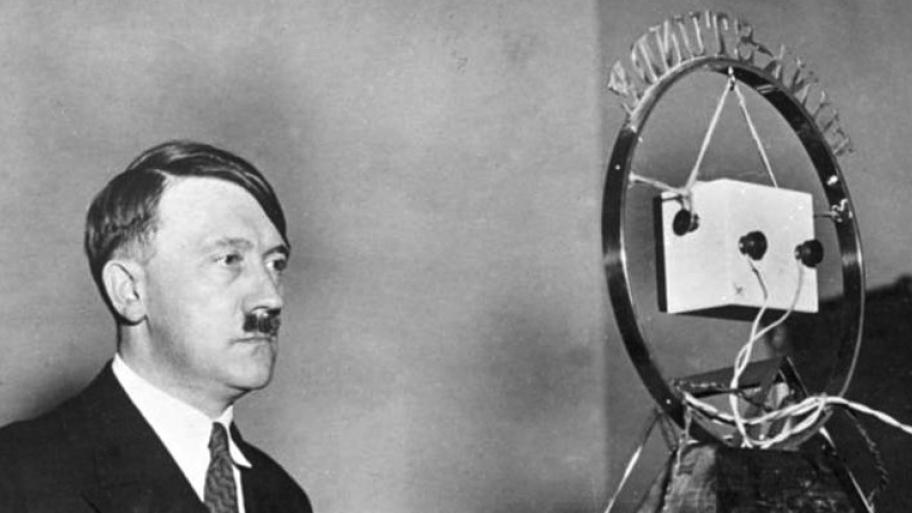 Adolf Hitler steht in dunklem Anzug, weißem Hemd und dunkler Krawatte vor einem Rundfunk-Mikrofon
