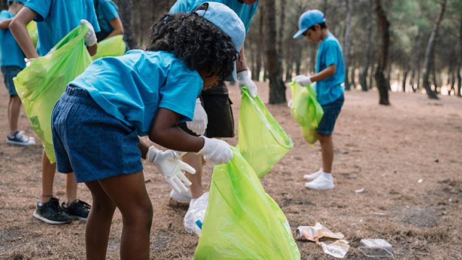 eine Gruppe Kinder sammelt Müll im Wald, sie tragen dieselben blauen Hemden und Basecaps und Handschuhe. Das Mädchen im Vordergrund bückt sich gerade