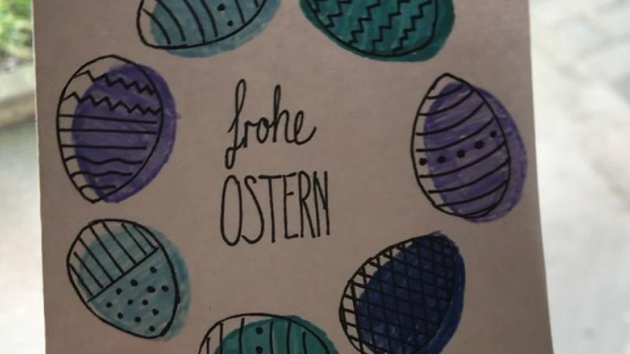 In der Mitte einer Osterkarte steht "frohe Ostern". Die Worte stehen in einem Kranz aus bunten Ostereiern.