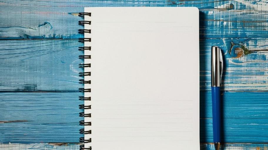 Foto von einem Notizbuch. Es liegt auf einem blauen Hintergrund aus Holz. Rechts daneben liegt ein blauer Kugelschreiber.