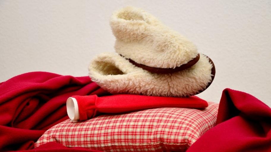 Es sind eine kuschelige Decke, ein Kissen, eine Wärmflasche und warme Hausschuhe übereinander gestapelt.