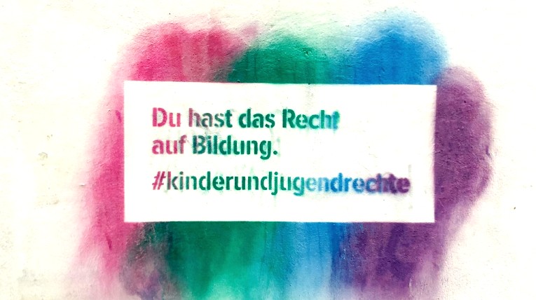 ein Graffiti an einer Hauswand mit dem Text: "Du hast das Recht auf Bildung #kinderundjugendrechte", der Hintergrund ist pink-grün-blau-lila gestreift