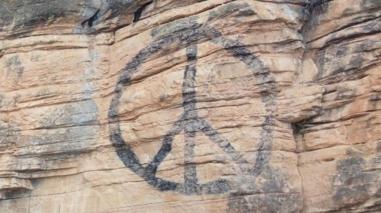 auf einer rötlichen Felswand ist in schwarz das bekannte, kreisförmige Peacesymbol aufgemalt