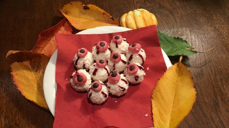 auf einem weißen Teller eine rote Papierserviette, darauf liegen 10 weiße Kokospralinen mit Augen verziert, drumherum Herbstlaub als Deko