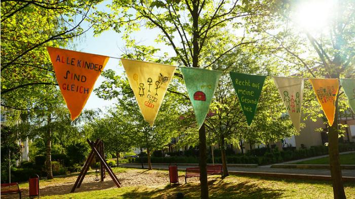 Wimpelkette aus 6 Wimpeln, die passend zu den Kinderrechten gestaltet sind. Im Hintergrund ist ein Spielplatz und Bäume.