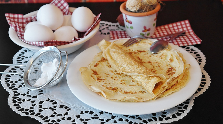 Eierkuchen liegen gestapelt auf einem Teller, daneben ein kleines Sieb mit Puderzucker und im Hintergrund eine Schale mit weißen Eiern