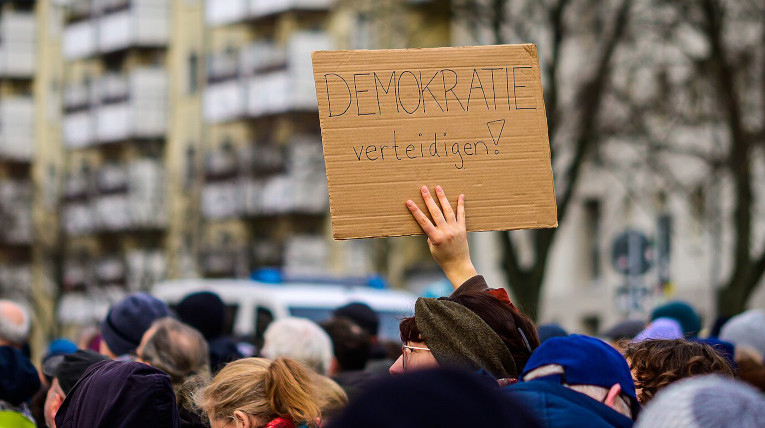 Eine Person hält ein Demoschild hoch auf dem steht "Demokratie verteidigen!", viele Menschen sind versammelt, um zusammen zu demonstrieren