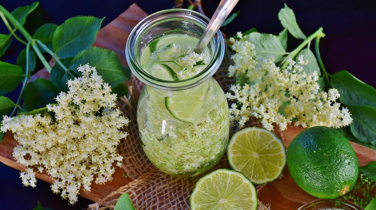 ein gedeckter Tisch, darauf eine Karaffe mit gelblicher Holunder-Limonade, daneben aufgeschnittene Limetten und weiße Holunderblüten