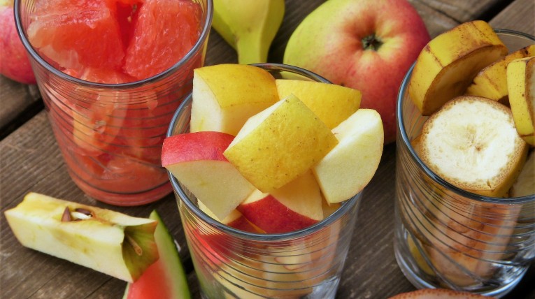 auf einem hölzernen Tisch stehen 3 Gläser gefüllt mit unterschiedlichen, geschnittenen Früchten, zum Beispiel Wassermelone, Apfel und Banane