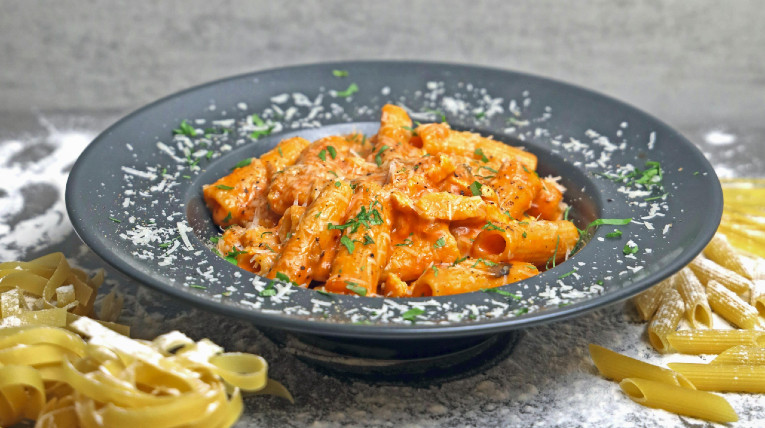 Dunkelgrauer Teller mit Nudeln in roter Soße. Darüber Parmesan und grüne Kräuter.