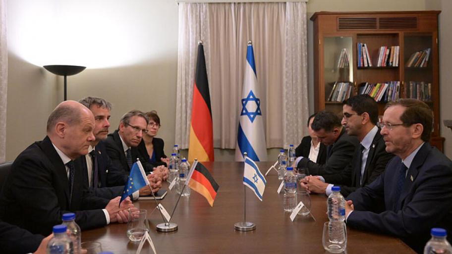 Acht Politiker und Politikerinnen sitzen sich gegenüber am Tisch. In der Mitte sieht man eine deutsche und eine israelische Flagge.