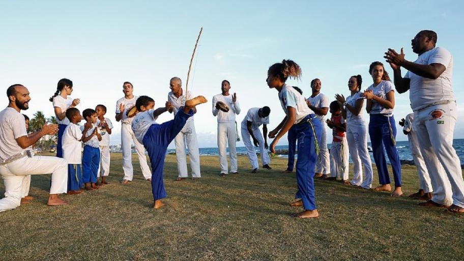eine Gruppe aus 16 Capoeira Tänzern (Männer, Frauen, Kinder) steht im Halbkreis auf einer Wiese um zwei Capoeira tanzende Kinder herum