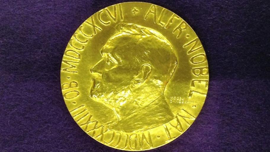 auf lila-farbenem Untergrund liegt die goldene Friedensnobelpreis-Medaille, darauf zu sehen das Porträt von Alfred Nobel in der Seitenansicht und eine Inschrift mit römischer Jahreszahl