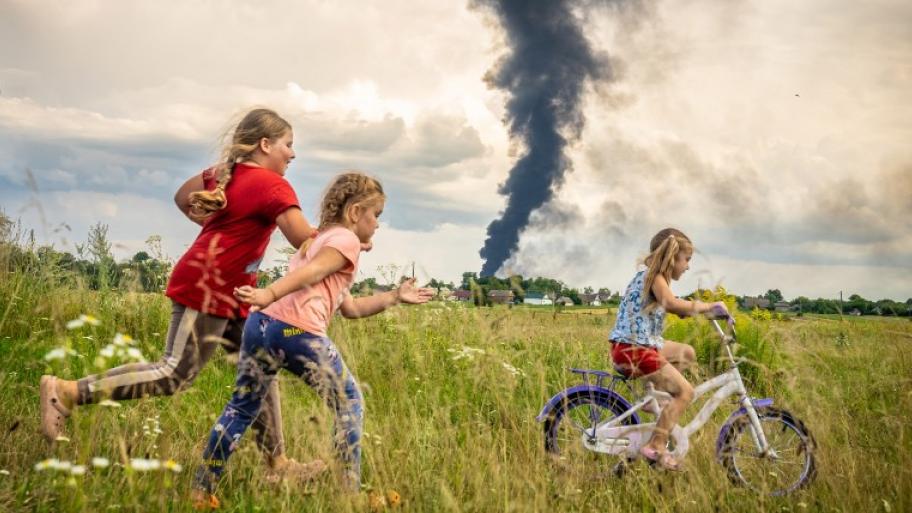 Drei Kinder sind zusehen, eins auf dem Fahrrad. im Hintergrund ist eine schwarze Rauchwolke.