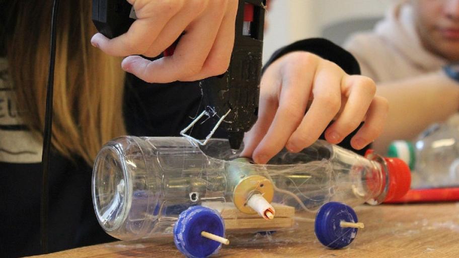 ein Kind basteltaus einer alten Plastikflasche und Deckeln mit der Heißklebepistole ein Auto