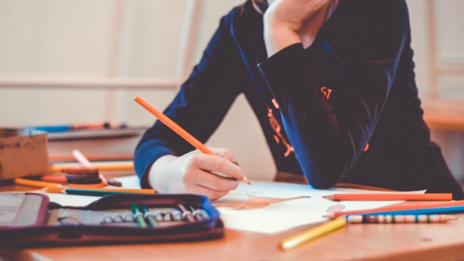 ein Kind sitzt an seinem Tisch im Klassenzimmer, vor ihm liegen Stifte, Federmappe, Arbeitsblätter ausgebreitet, es hält einen Buntstift in der Hand