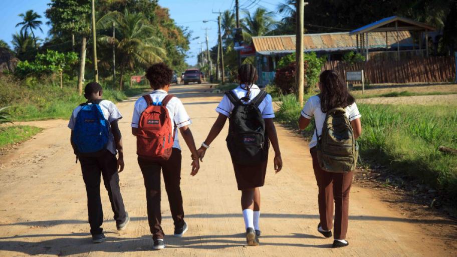 Vier Kinder gehen auf einer Straße zu Fuß in die Schule, sie  tragen Rucksäcke auf dem Rücken, zwei Kinder halten sich an der Hand
