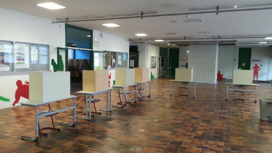 ein leeres Wahllokal im Vorraum einer Schule mit 6 Wahlkabinen, jeweils ein Tisch und ein Stuhl pro Kabine, rechts die Wahlurne
