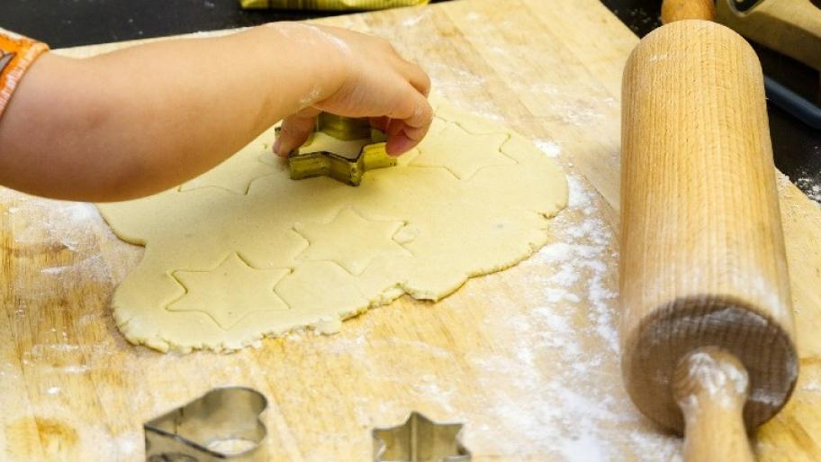 auf einer hölzernen Unterlage sticht eine Kinderhand aus ausgerolltem, hellem Plätzchenteig Sternchenplätzchen aus, daneben weiter Ausstechformen, ein Nudelholz, Mehlstaub