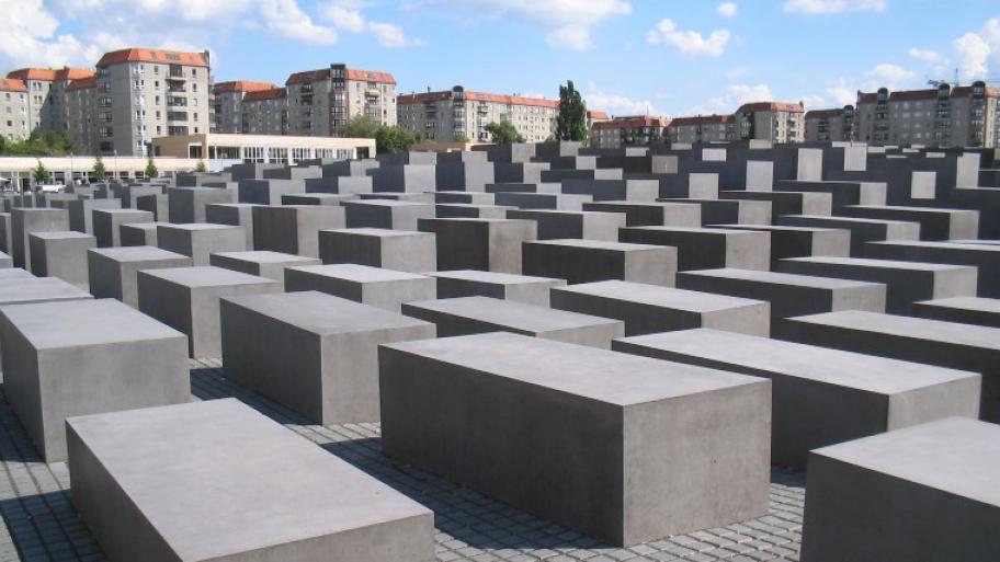 das Holocaust-Mahnmal in Berlin, eine große Fläche voll mit grauen Betonstelen, unterschiedlich hoch, im Hintergrund Häuser