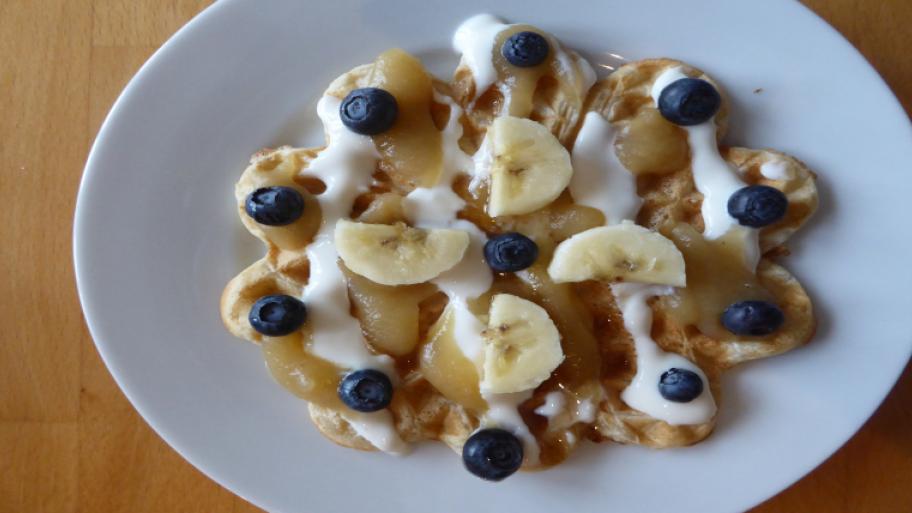 auf einem weißen Teller eine runde Waffel, garniert mit Bananenscheiben, Blaubeeren und Joghurt