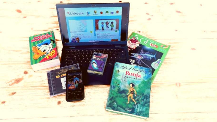 auf hellem Holzboden liegen und stehen ein aufgeklappter Laptop mit der Seitenansicht von kindersache, eine drei Fragezeichen Kassette, eine Ausgabe vom Kindermagazin Geolino, ein Donald Duck Comic, eine CD von Gregs Tagebuch