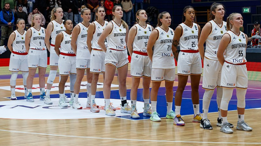 die Spielerinnen der deutschen Basketball-Nationalmannschaft 2022 stehen in weißem Nationaltrikot aufgereiht hintereinander auf dem Basketballfeld, im Hintergrund Zuschauer*innen
