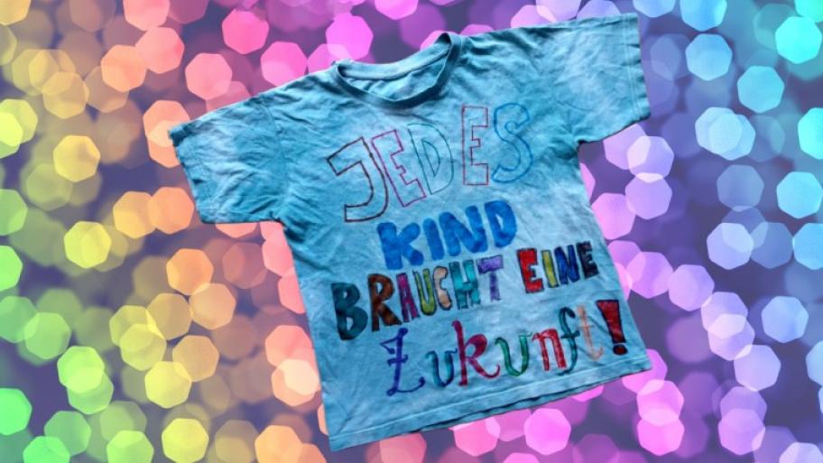 ein türkises T-Shirt, auf der Vorderseite steht in bunter Schrift mit Stoffmalstiften geschrieben "Jedes Kind braucht eine Zukunft!"