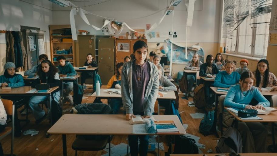 Ein Klassenzimmer ist abgebildet. Es sitzen Schüler und Schülerinnen in dreier Reihen an ihren Tischen. In der Mitte steht ein Mädchen hinter ihrem Tisch und stützt sich darauf ab.