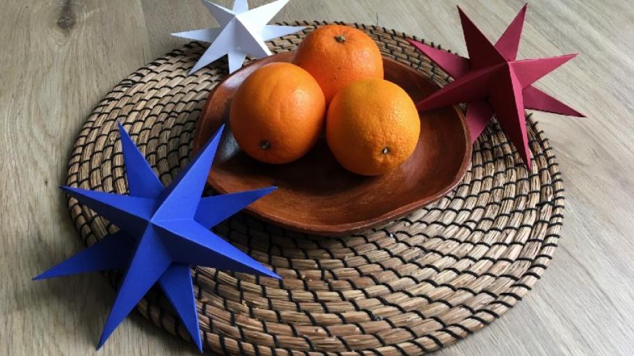 3D Sterne aus Papier in blau, rot und weiß; in der Mitte des Tisches befindet sich auf einem Korbuntersetzer ein Holzteller mit drei Orangen