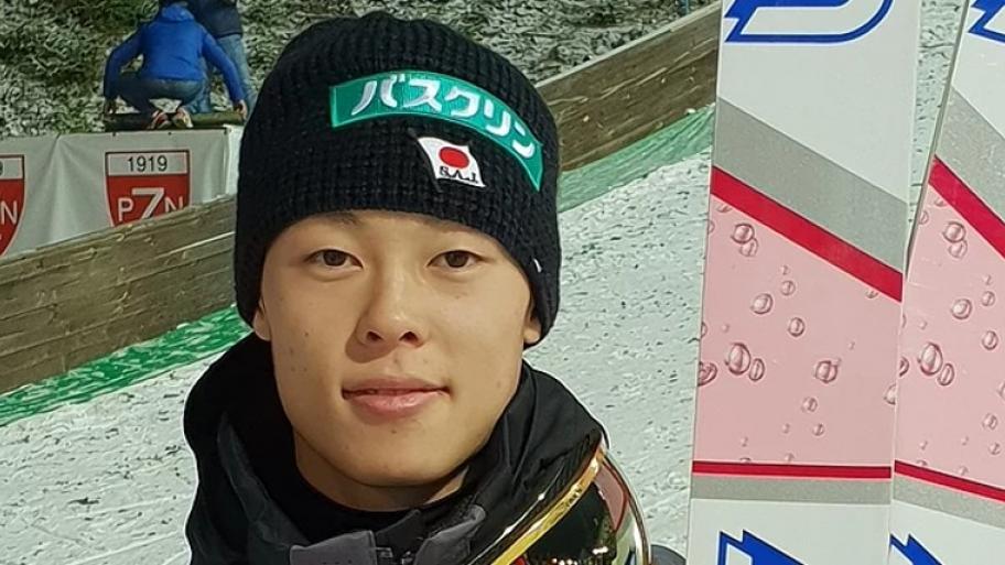 Großaufnahme von dem Gesicht eines japanischen Skispringers. Er trägt eine schwarze Mütze und hält links von sich, seine Skier aufrecht.