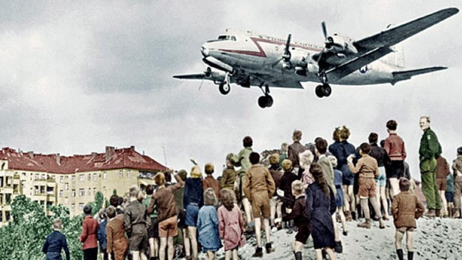 ein "Rosinenbomber" während der Berliner Luftbrücke 1948 im Landeanflug, darunter viele Kinder auf einem Schuttberg stehend, im Hintergrund Häuser. darunte