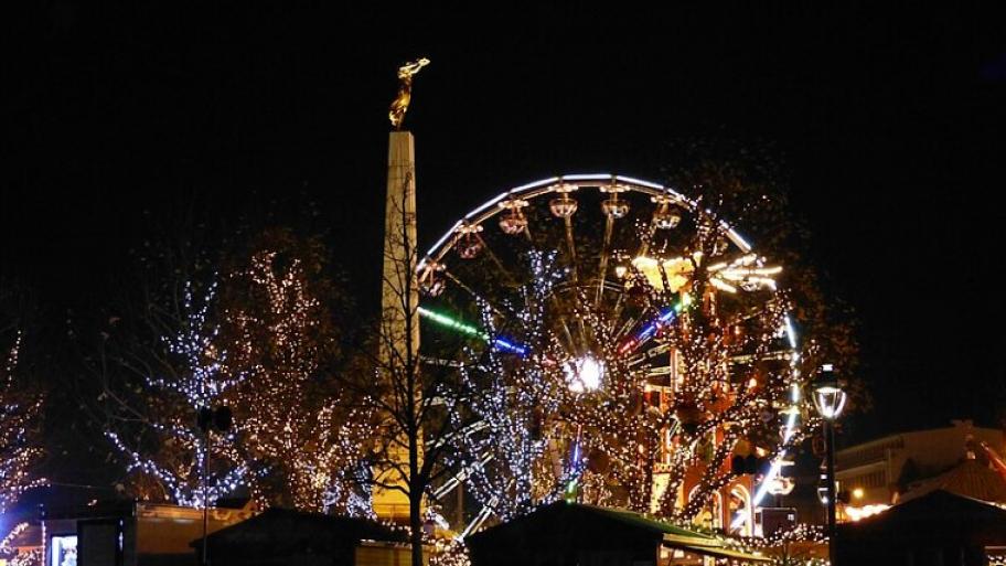 Zu sehen ist der Konstitutionsplatz in Luxemburg-Stadt mit der Statur der goldenen Frau am Abend. Auf dem Platz stehen beleuchtete Bäume, Weihnachtsmarkthütten und ein Riesenrad.