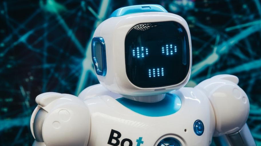der Oberkörper eines weißen Roboters, auf der Brust steht Bot, sein Gesicht ist eine digitale Anzeige, der dunkle Hintergrund ist durchzogen von mehreren
