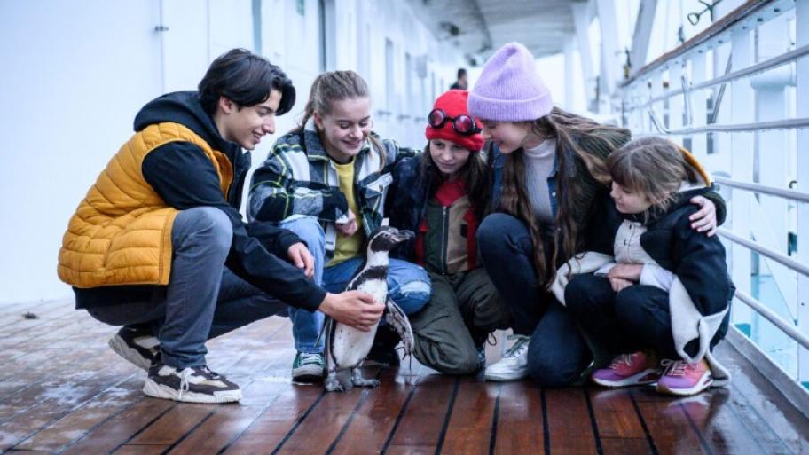 Auf dem Bild sind die 4 Schwestern, die den Pinguin umarmen. Daneben ist ein Junge mit einer gelben Weste. Alle sind glücklich und sie befinden sich auf einem Boot.