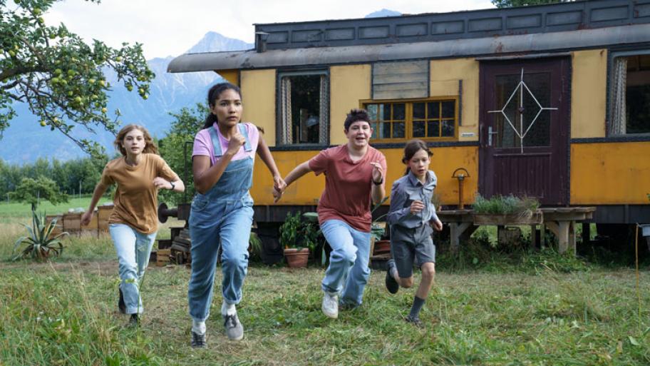 Vier Kinder rennen auf einer Wiese, zwei Mädchen und zwei Jungen; im Hintergrund szeht ein alter Bauwagen