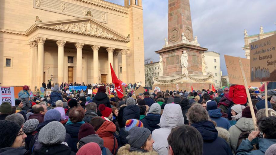 Eine Menschenmenge, einige mit Fahnen und Schildern. Im Hintergrund sieht man auf den Treppen einer Kirche viele Menschen mit Plakaten