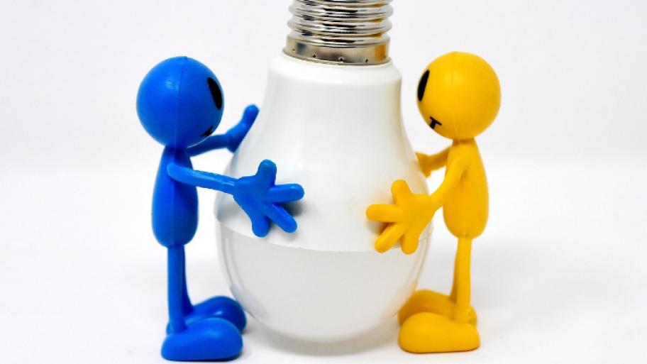 Ein blaues und ein gelbes Männchen halten eine Energiesparlampe gemeinsam (Glühbirne) in ihren Händen