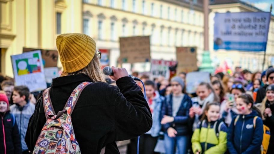 ein Klimastreik von Fridays for Future: im Vordergrund ein junger Mensch mit gelber Mütze, dunkler Jacke, buntem Rucksack und Mikrofon in der Hand von hinten, davor eine große Menge junger demonstrierender Menschen