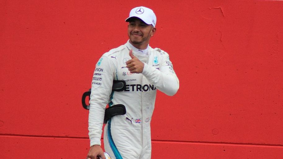 der Formel 1 Rennfahrer Lewis Hamilton im weißen Rennanzug, Helm in der rechten Hand, streckt den linken Daumen in die Luft