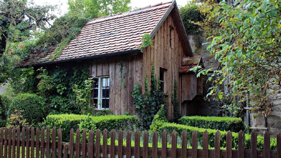 ein mysteriös aussehendes kleines Häuschen aus dunklem Holz ist in einem verwachsenen Garten mit Gartenzaun ist zu sehen