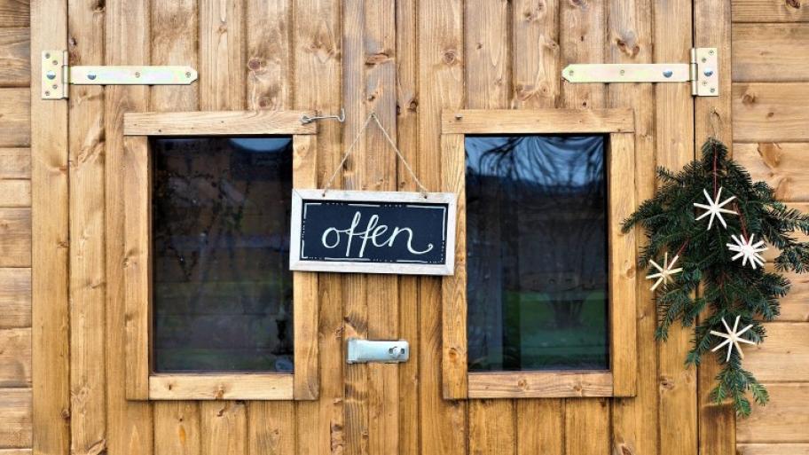 Eingang eines Holzhauses an dem ein Schild hängt mit der Aufschrift: "offen; rechts daneben hängt ein Tannenzweig