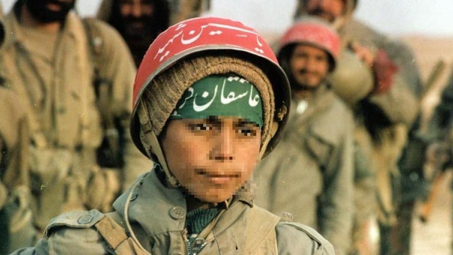 ein Junge in iolivgrüner Soldatenuniform mit Stahlhelm auf dem Kopf, auf Helm und Stirn arabische Schriftzeichen, im Hintergrund erwachsene, männliche Soldaten 