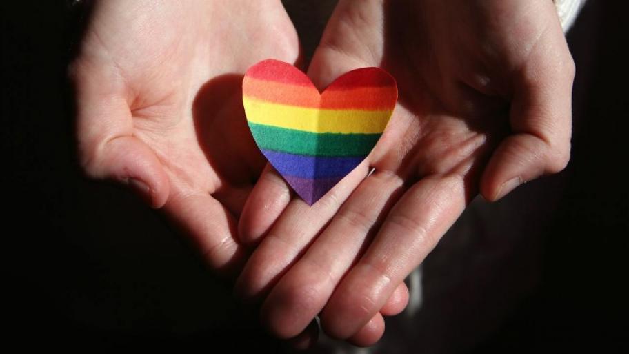 Zwei Hände halten ein ausgeschnittenes Herz. Auf dem Herz sind die Farben der LGBTQ+ Flagge