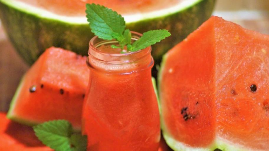 eine Wassermelone in mehrere Teile aufgeschnitten, in der Mitte vorn ein Glas mit Melonen-Bowle mit frischer Minze