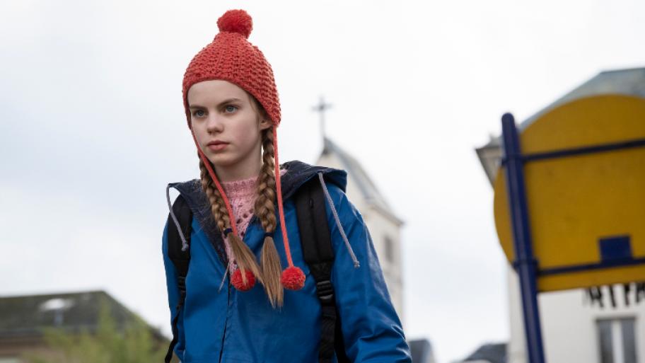 ein blondes Mädchen um die 14 Jahre trägt eine rote Bommmelmütze, eine blaue Regenjacke und guckt links an einwm Punkt etwas abseits von der Kamera