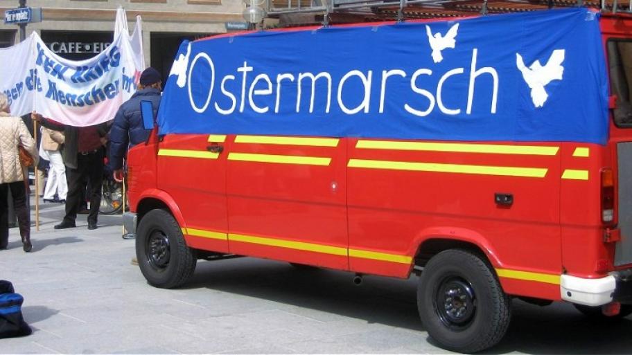 an einem roten Kleintransporter hängt ein großes blaues Transparent, darauf in weiß die Aufschrift "Ostermarsch" und drei weiße Friedenstauben. im Hintergrund ein paar Demonstrierende