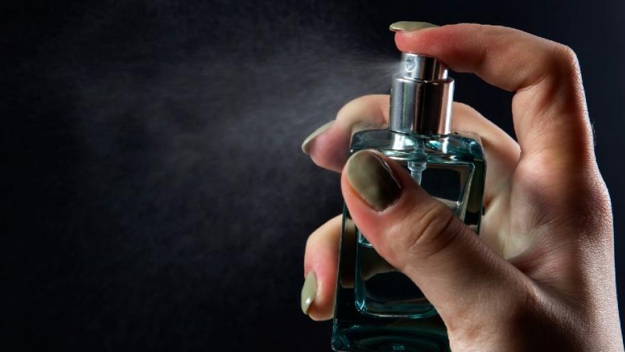 Eine Hand mit Nagellack auf den Fingernägeln sprüht Parfüm. Dahinter ist ein schwarzer Hintergrund.