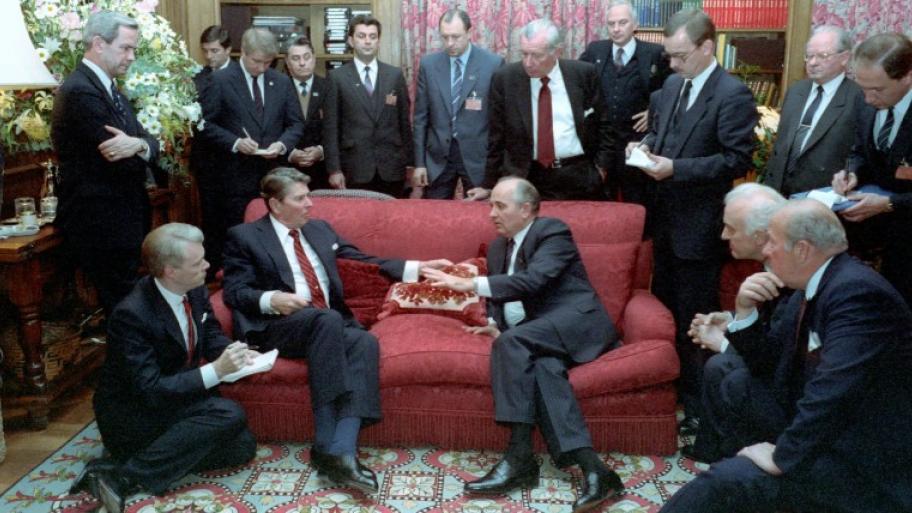 die Präsidenten der USA und der Sowjetunion Ronald Reagan und Michael Gorbatschow (beide sitzend auf einem roten Sofa) treffen sich zum Gespräch, drumherum etliche Vertreter beider Länder, alle tragen dunklen Anzug und Krawatte 
