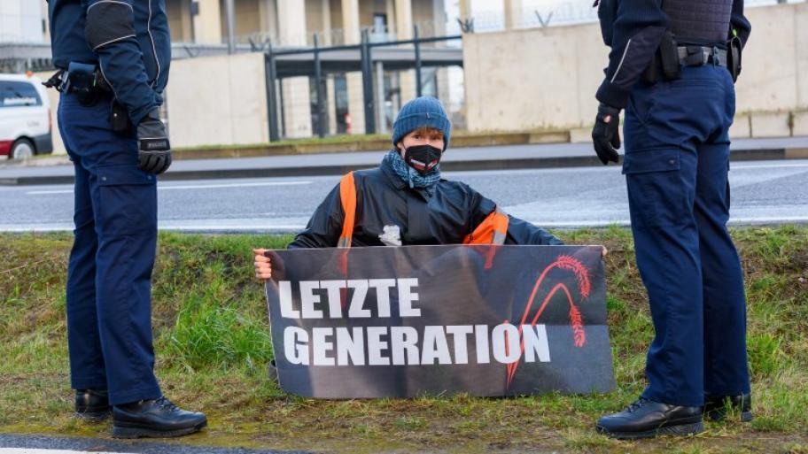 ein Mitglied der „Letzten Generation” sitzt bei der Blockade des Berliner Flughafens im Februar 2022 auf einem Rasenstreifen mit einem Transparent in Händen auf dem "Letzte Generation" steht, links und rechts steht jeweils ein Polizist in Uniform 